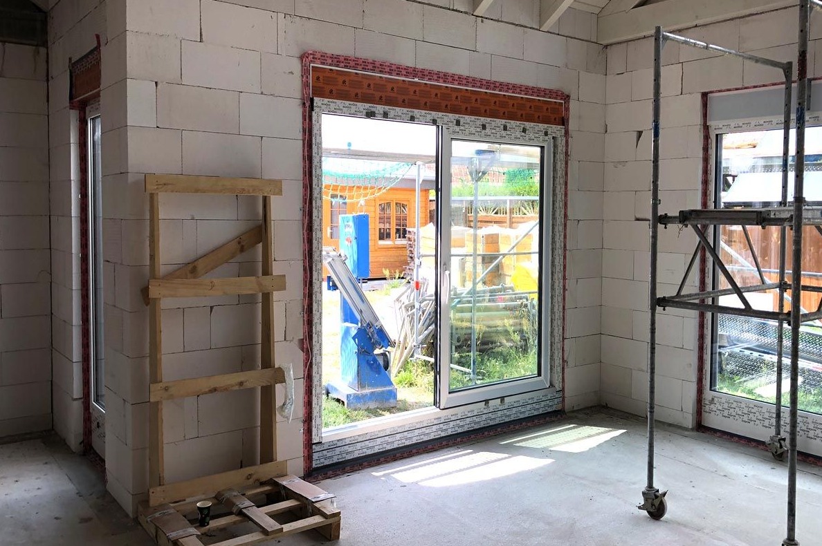 Wnętrze w trakcie budowy z widocznymi oknami PCV, rusztowaniem i pustakami z betonu komórkowego, z maszyną do montażu okien widoczną przez otwarte okno.