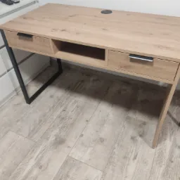 Biurko z blatem imitującym jasne drewno, z dwoma szufladami i otwartą półką, na metalowych nogach w kształcie litery 'U', widoczne na tle białej komody i podłogi z paneli.