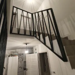 Nowoczesna balustrada schodowa z czarnych, ukośnych prętów, widok z dołu, z fragmentem kuchni w tle.