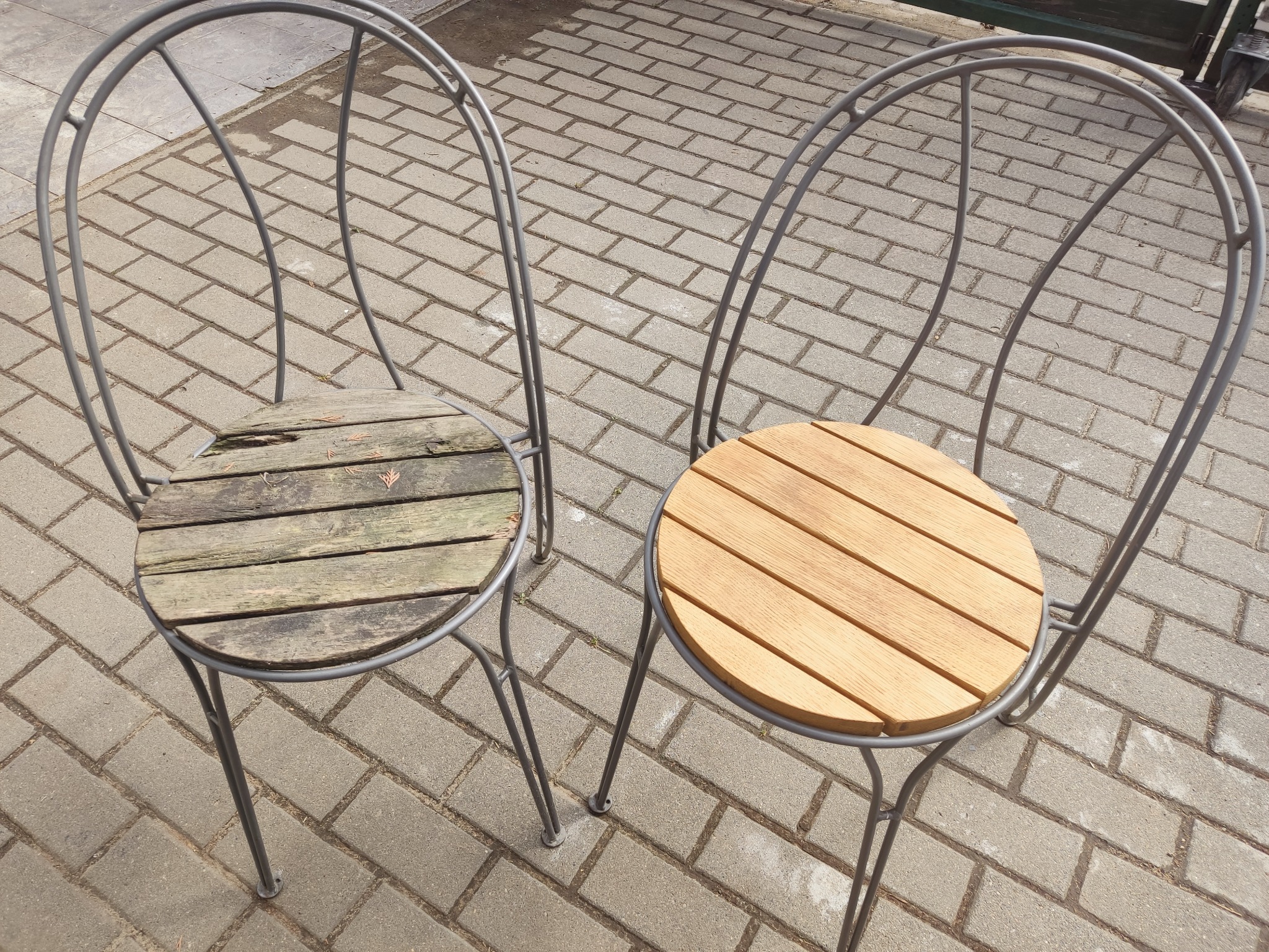Dwa metalowe krzesła ogrodowe z drewnianymi siedziskami: jedno z nowym, jasnym drewnem, drugie ze starym, zniszczonym drewnem, na tle kostki brukowej.