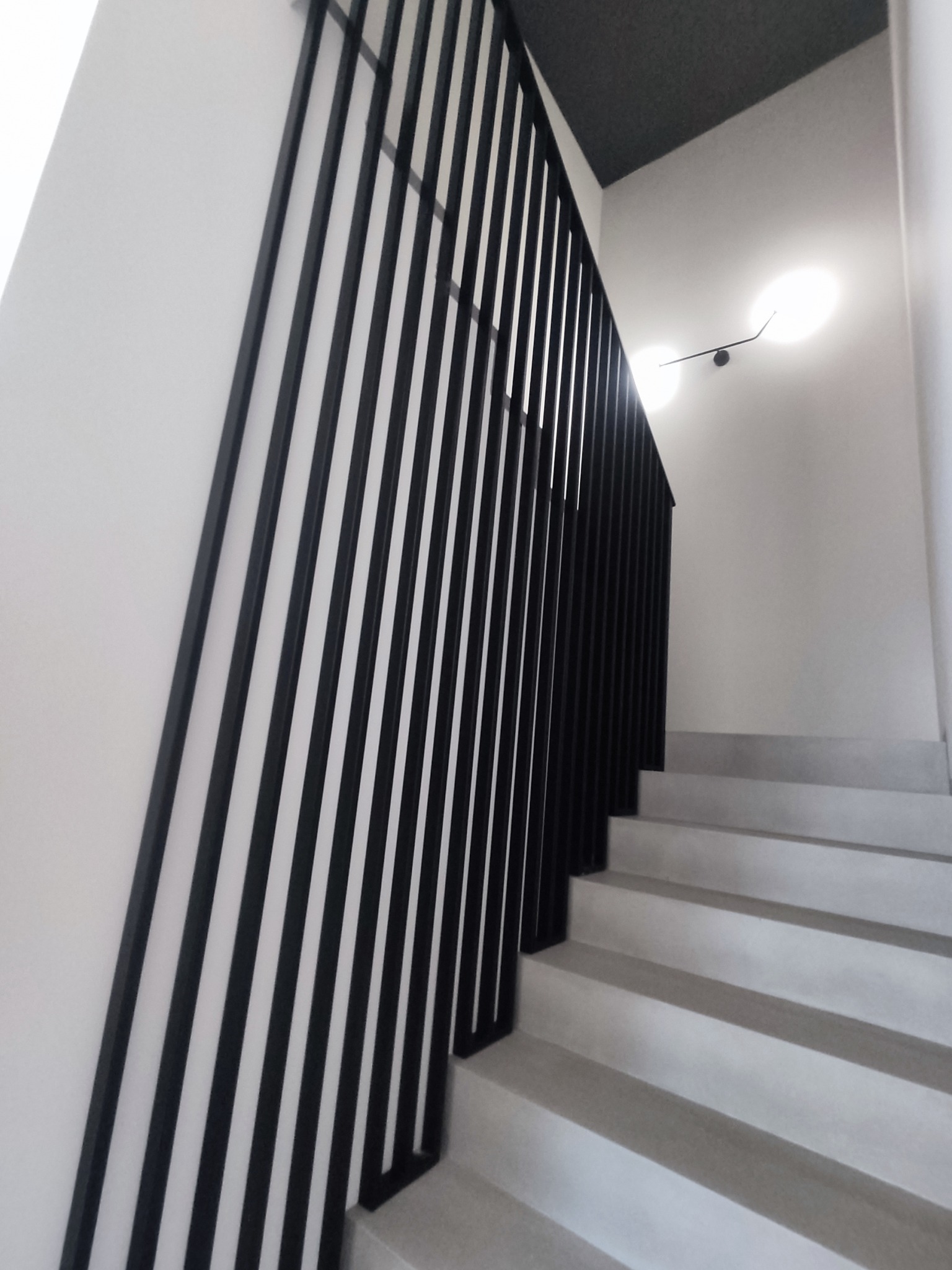 Nowoczesna balustrada schodowa z czarnych, pionowych profili na tle białych ścian i betonowych schodów, oświetlona minimalistyczną lampą sufitową.