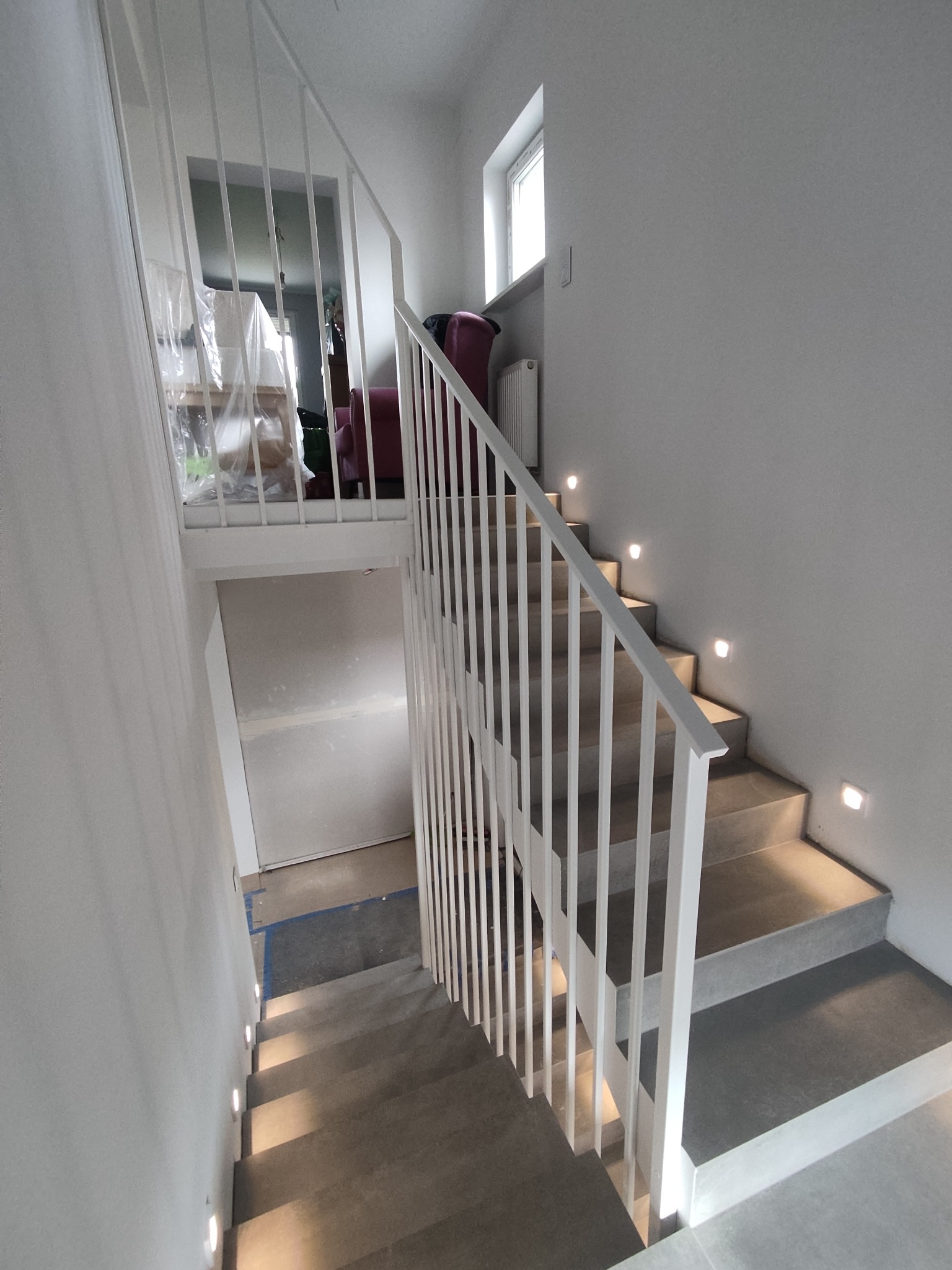 Nowoczesne schody z szarymi stopniami i białą, minimalistyczną balustradą, oświetlone punktowymi lampkami w ścianie, widok z góry.