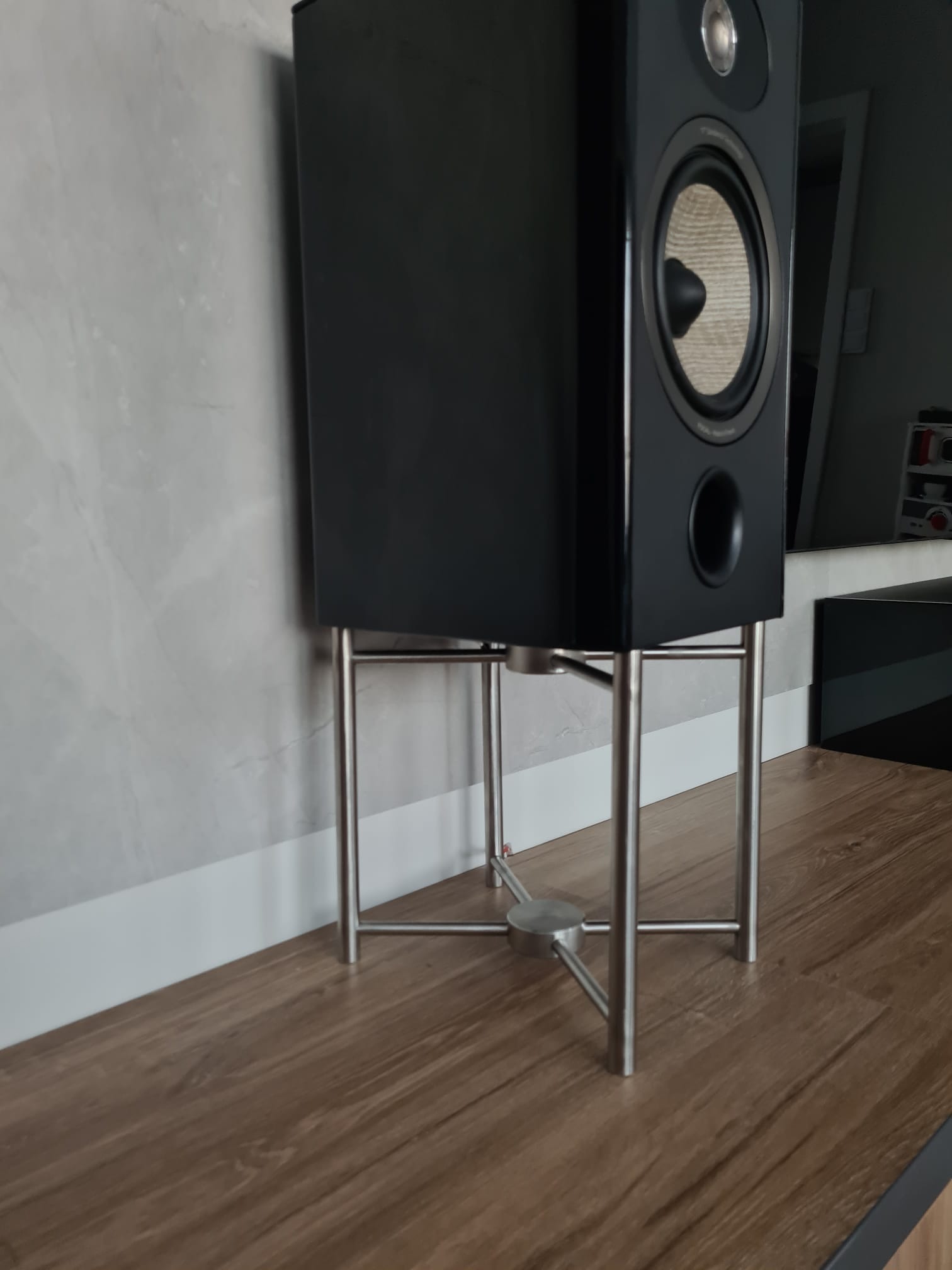 Czarna kolumna głośnikowa na metalowym stojaku, ustawiona na podłodze imitującej drewno, przy szarej ścianie z białą listwą przypodłogową.