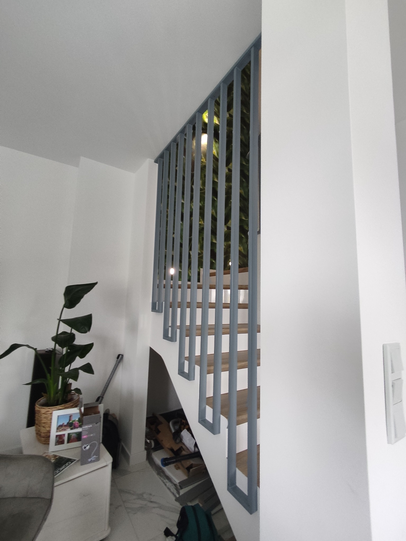 Nowoczesne schody z drewnianymi stopniami i szarą balustradą w jasnym wnętrzu. W tle widoczna zieleń roślin, a obok schodów, na komodzie, stoi roślina doniczkowa.