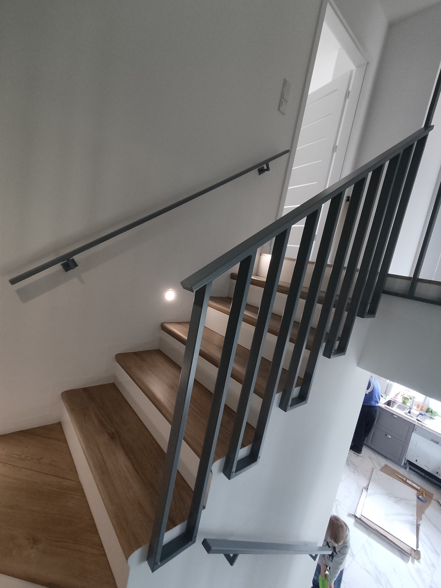 Nowoczesne schody drewniane z szarą metalową balustradą, widok z dołu, w tle fragment kuchni i osoba dorosła oraz dziecko.