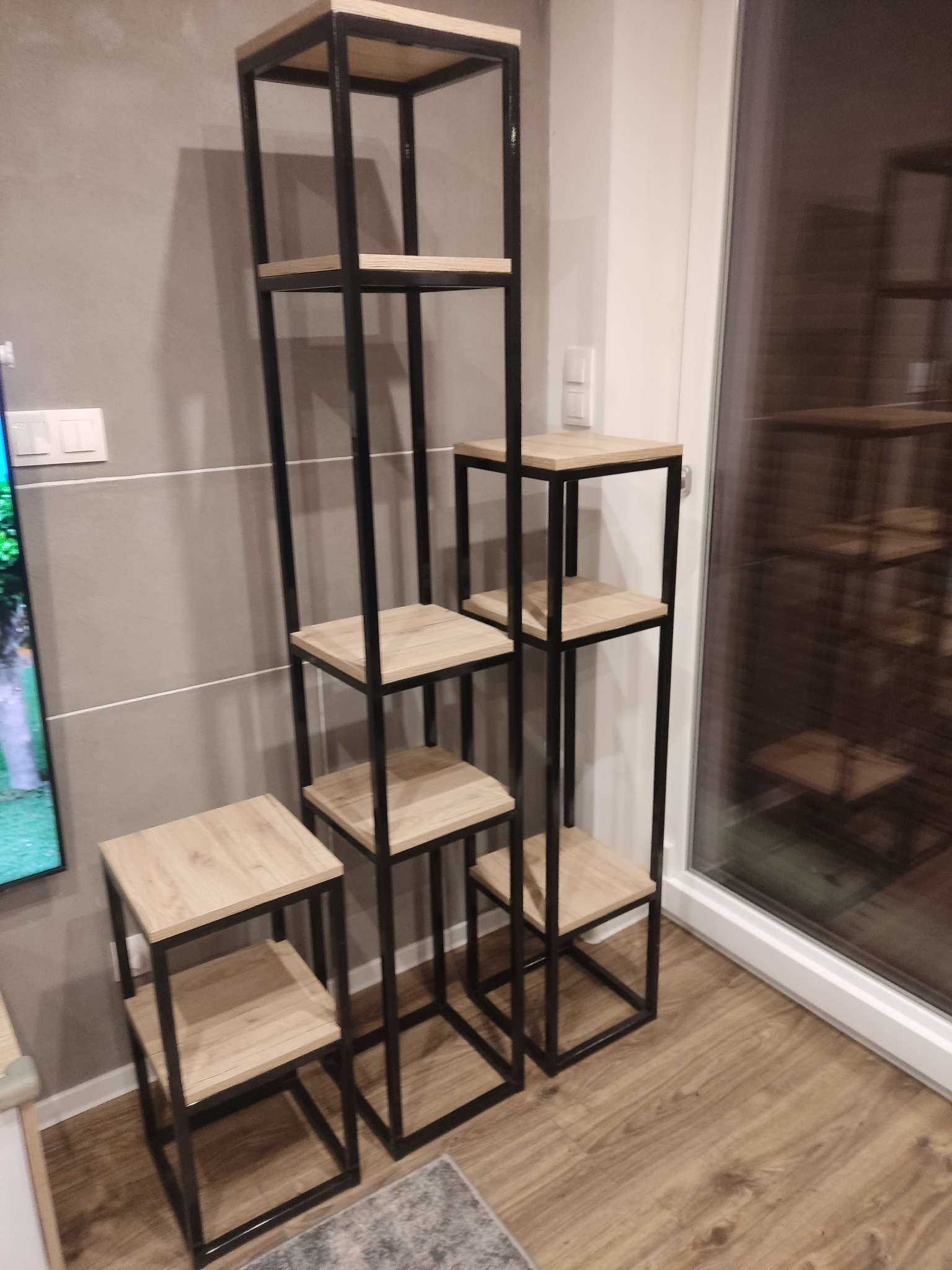 Regał modułowy z drewnianymi półkami i czarną metalową ramą, ustawiony schodkowo w nowoczesnym wnętrzu z panelami podłogowymi i szarą ścianą.