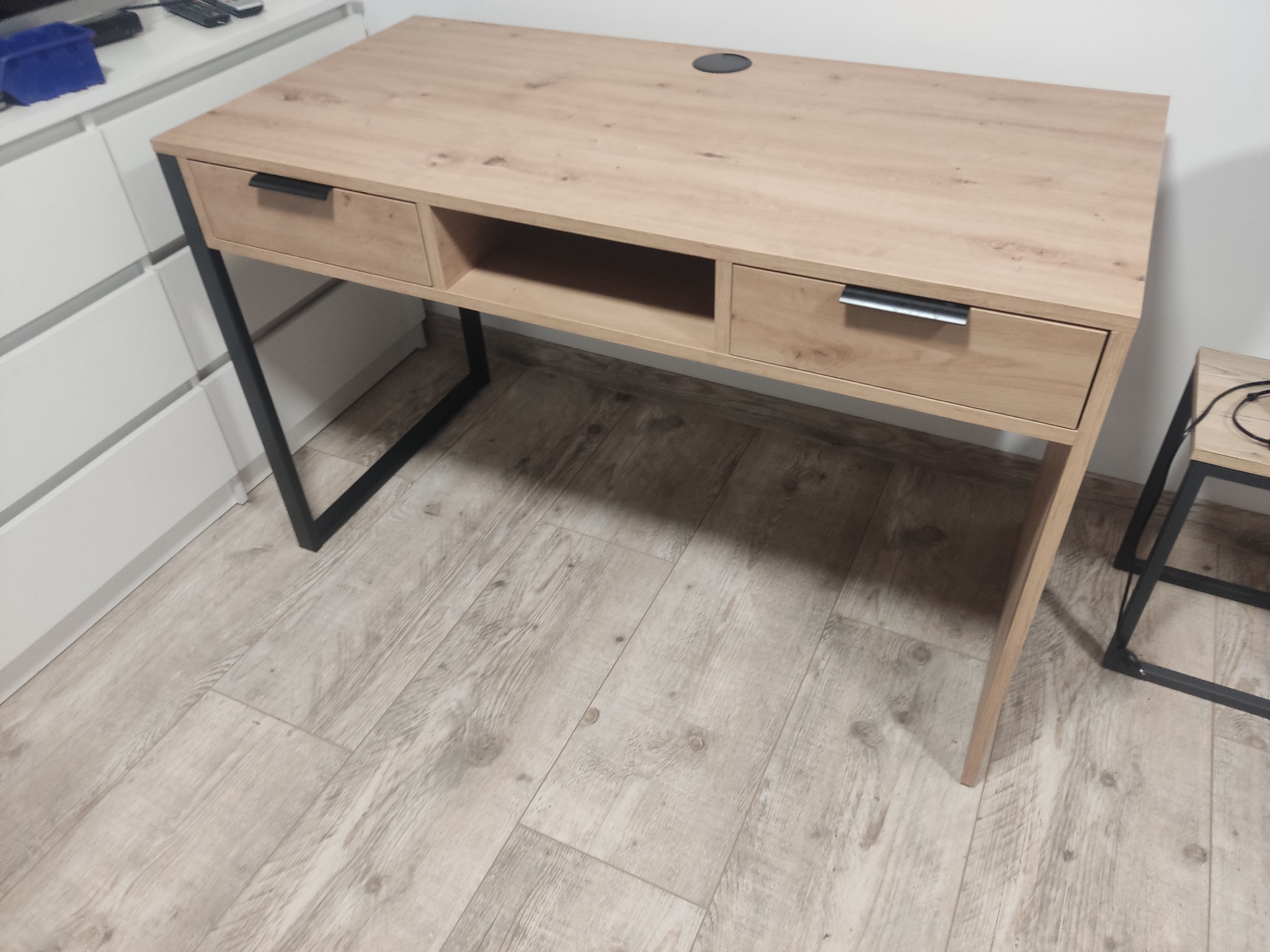 Biurko z blatem imitującym jasne drewno, z dwoma szufladami i otwartą półką, na metalowych nogach w kształcie litery 'U', widoczne na tle białej komody i podłogi z paneli.