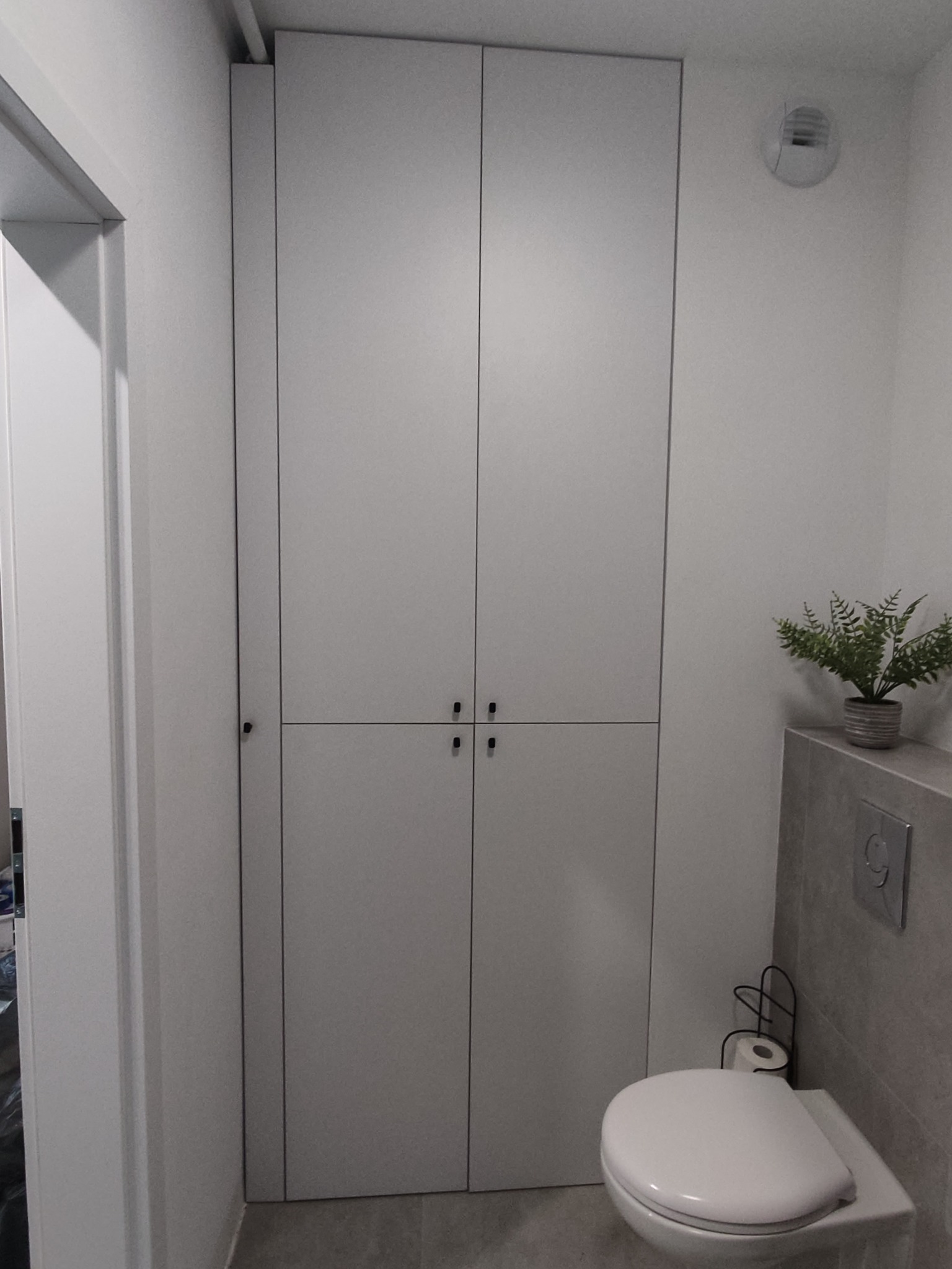 Biała, wysoka szafa z czterema drzwiami i minimalistycznymi uchwytami w łazience, obok toaleta wisząca i roślina w doniczce na zabudowie stelaża.