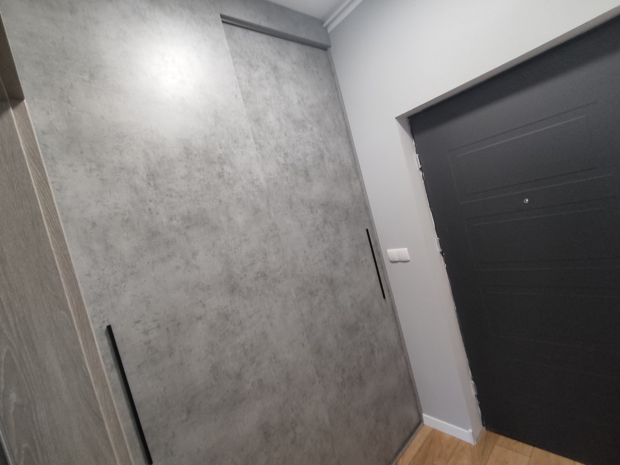 Nowoczesna szafa wnękowa z przesuwnymi drzwiami o szarej, betonowej fakturze, z minimalistycznymi, czarnymi uchwytami, przylegająca do ściany obok czarnych drzwi wejściowych.