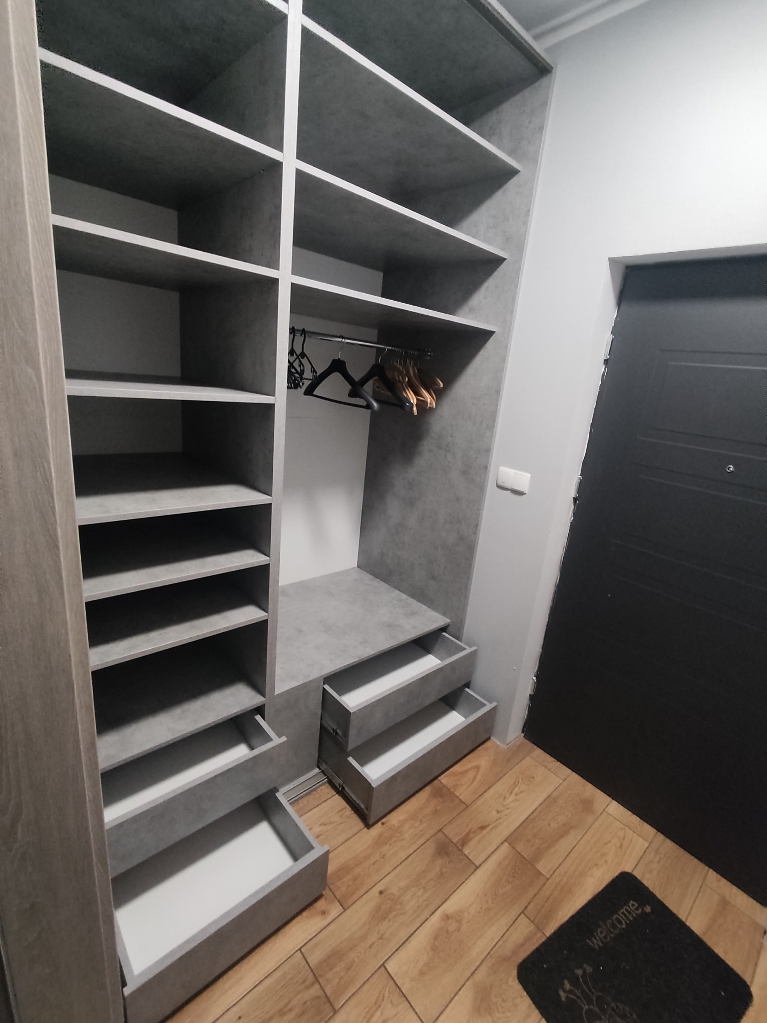 Szara szafa wnękowa z otwartymi półkami i szufladami, widoczne wieszaki na ubrania, na podłodze mata powitalna.