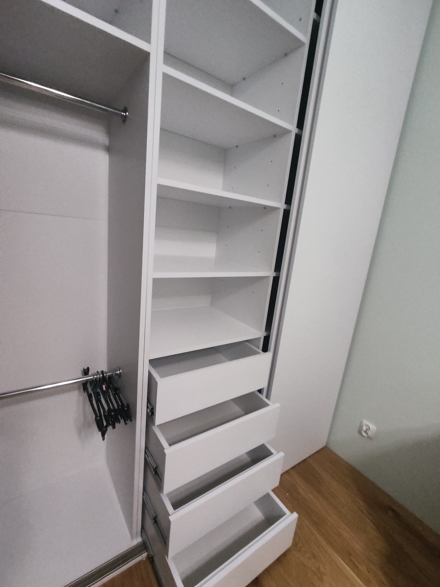 Wnętrze białej szafy wnękowej z otwartymi szufladami i półkami, widoczne drążki na ubrania i wieszaki.
