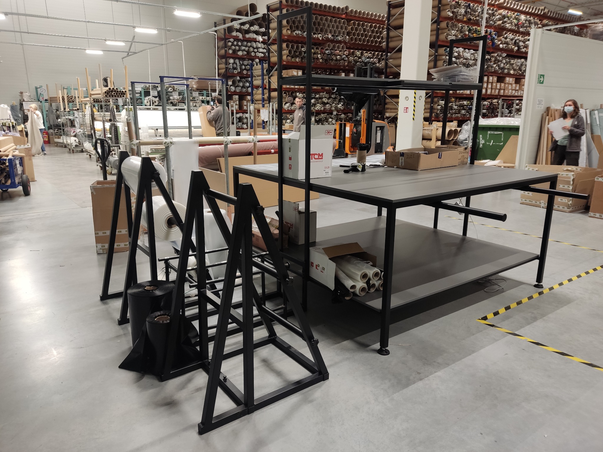 Wnętrze hali produkcyjnej z regałami wypełnionymi rolkami materiałów, stanowisko pracy z metalowym stołem i stojakiem na rolki folii, osoba w maseczce w tle.