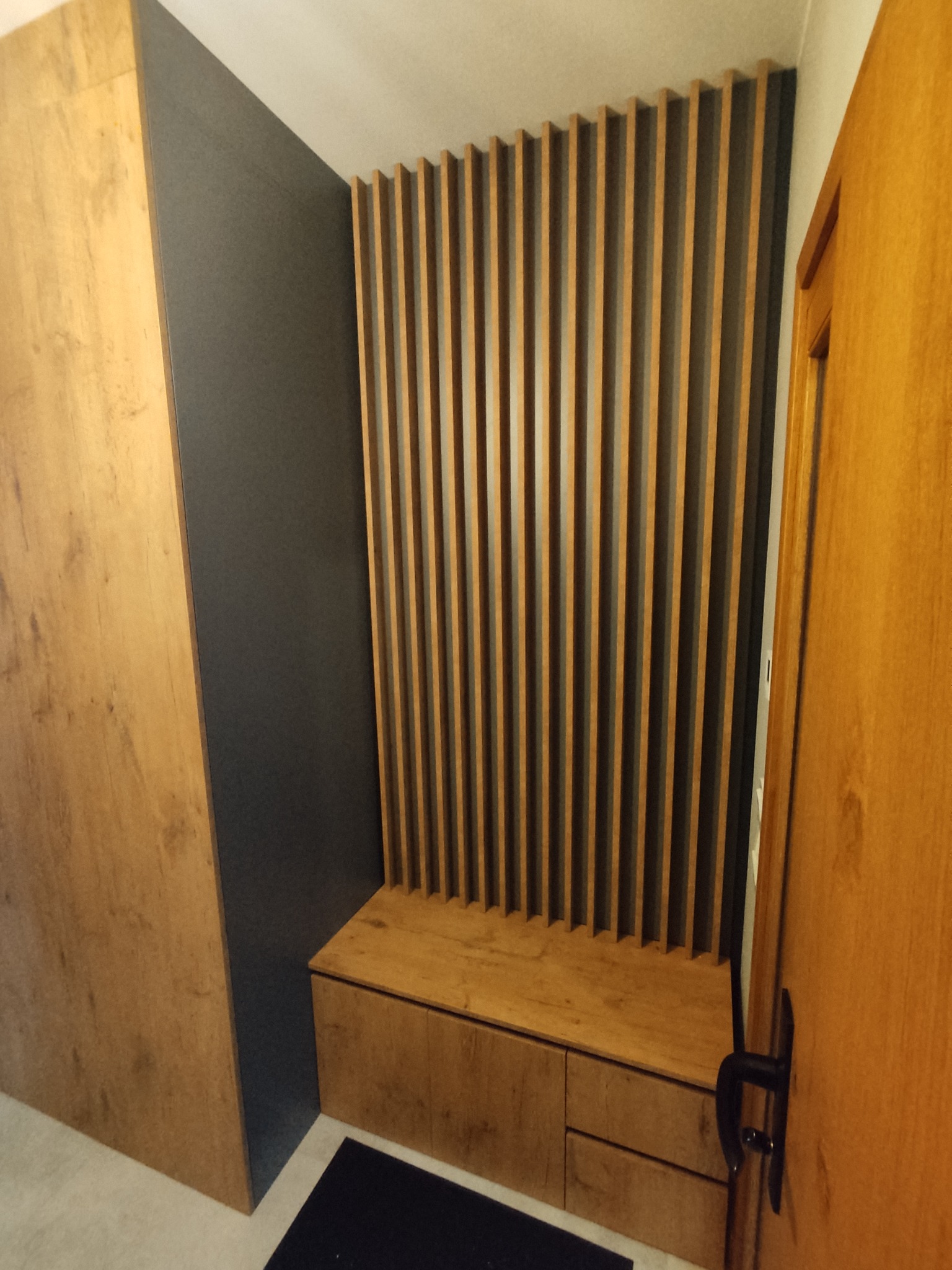 Wnęka z szafą i siedziskiem z drewnianymi lamelami na ścianie, szara ściana, widoczna drewniana podłoga.