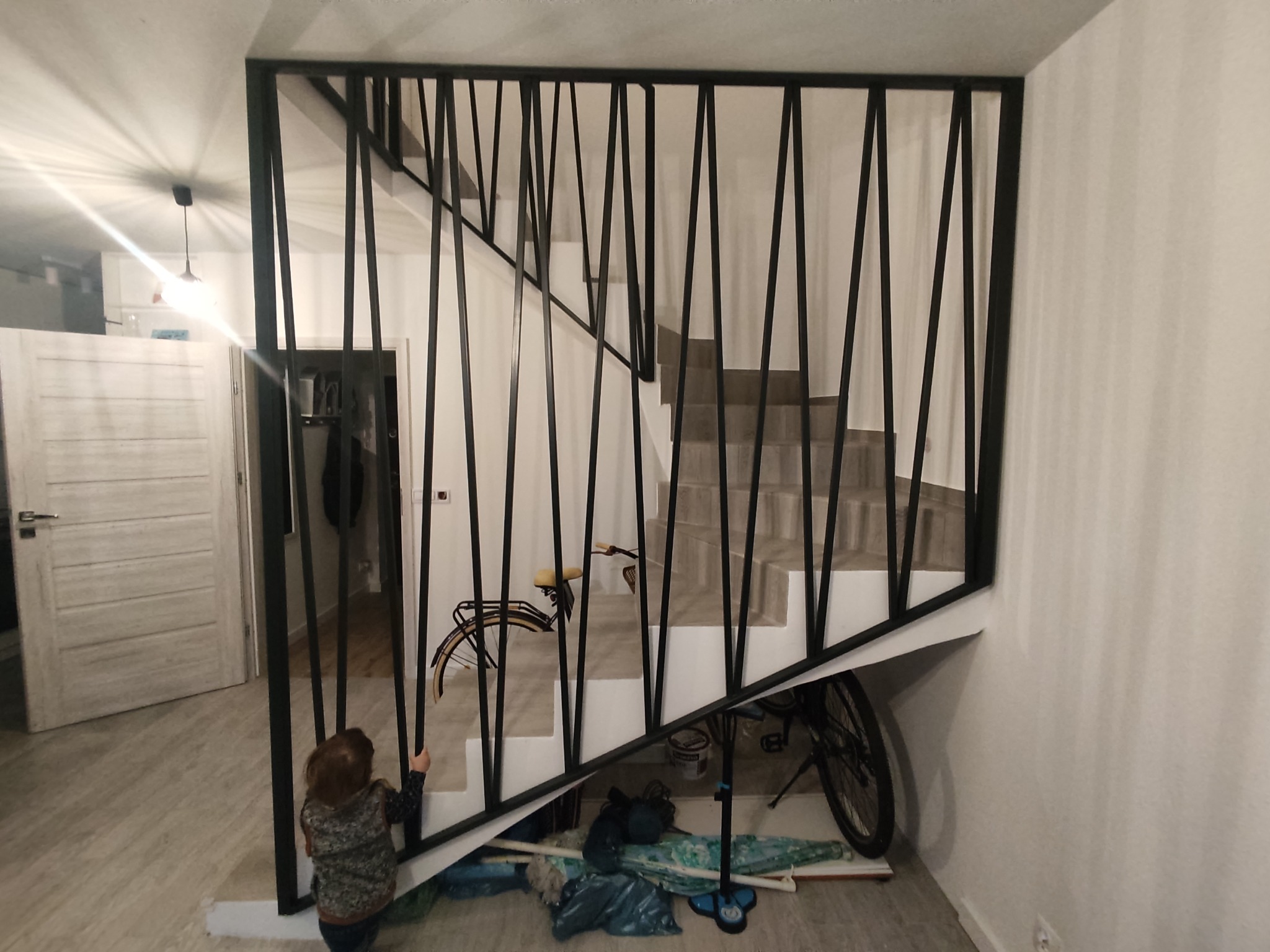 Nowoczesna balustrada schodowa z czarnych, pionowych prętów o nieregularnym rozstawie, widok na schody z szarymi stopniami, w tle rower i bałagan, mała dziewczynka trzyma się balustrady.