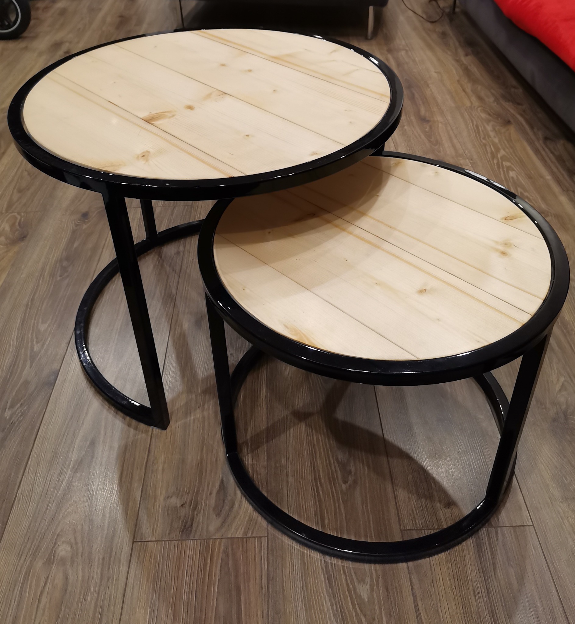 Dwa okrągłe stoliki kawowe z jasnego drewna z czarnymi metalowymi nogami, ustawione jeden na drugim na drewnianej podłodze.