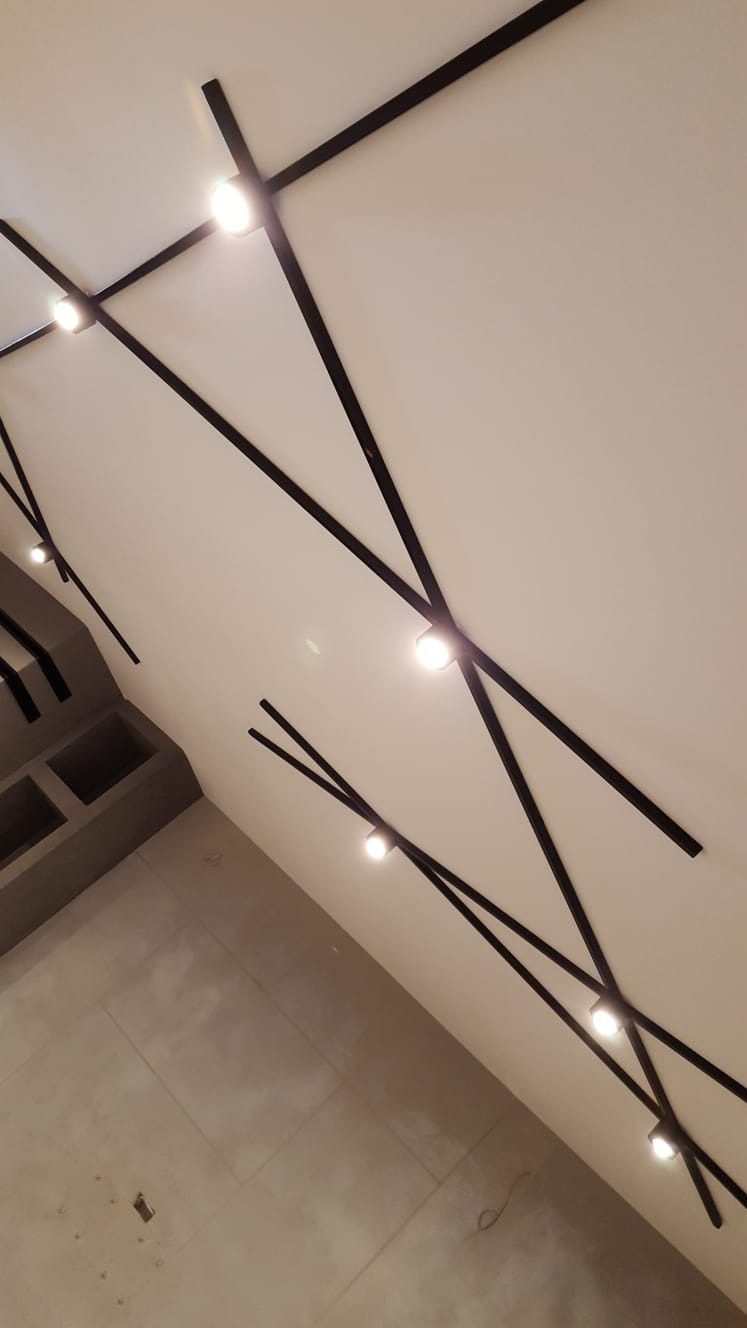 Nowoczesny sufit z geometrycznym układem czarnych listew i punktowym oświetleniem LED, widok z dołu na szary betonowy element zabudowy ściany.