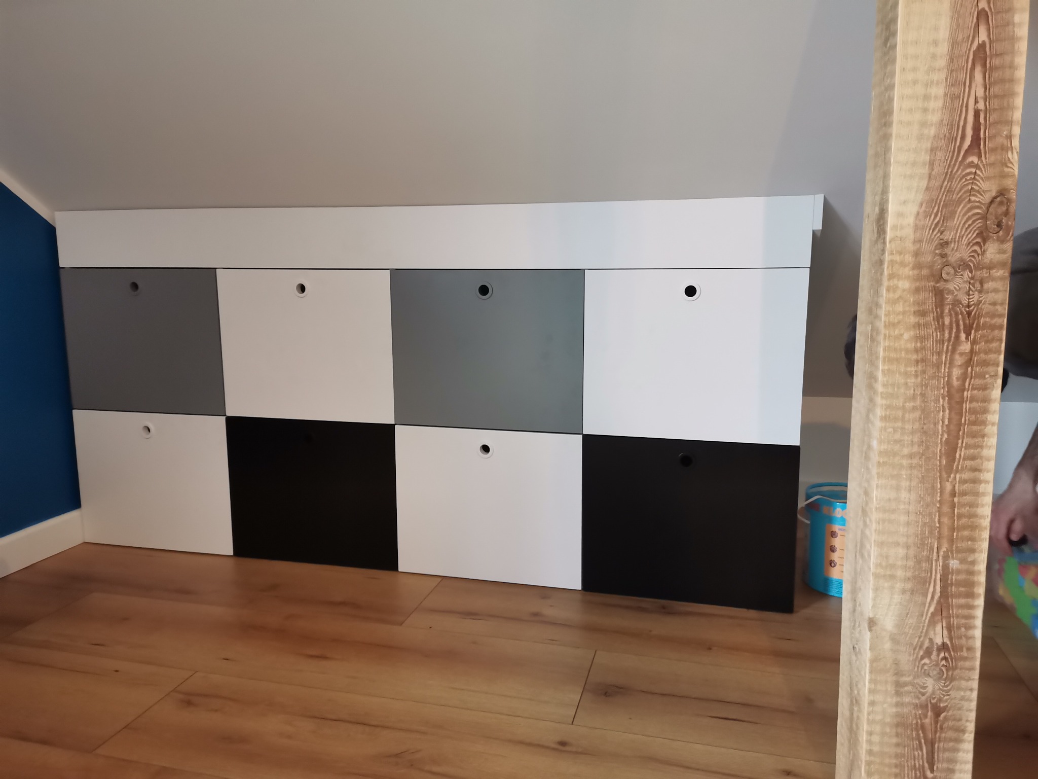 Zabudowa szafkowa z frontami w kolorach białym, szarym i czarnym, umieszczona pod skosem dachu na poddaszu, widok z niskiej perspektywy, z boku widoczny fragment drewnianej belki.