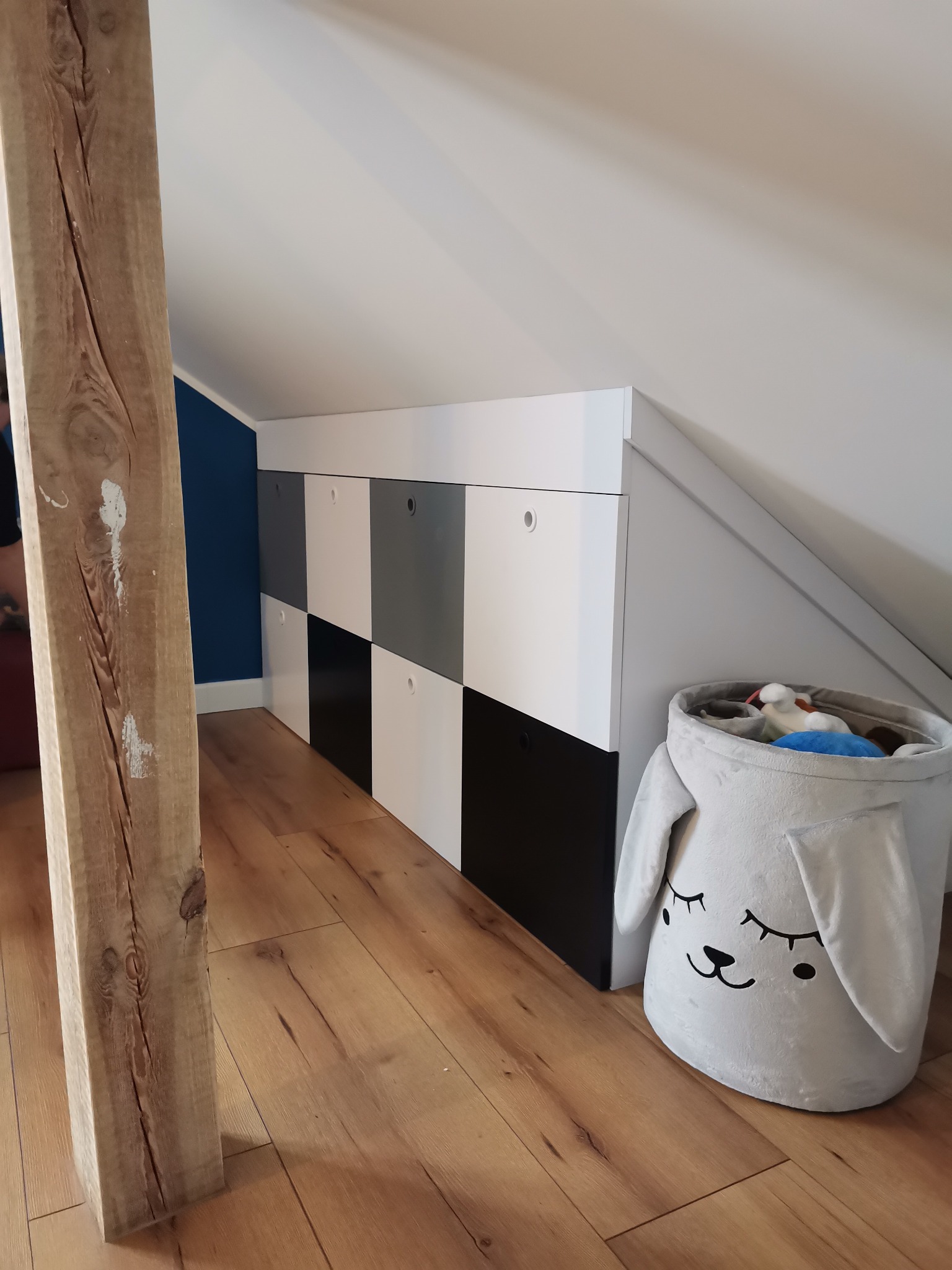 Szafa wnękowa na poddaszu w szachownicę (biało-szaro-czarna) z okrągłymi uchwytami, obok kosz na zabawki w kształcie królika, widoczna belka konstrukcyjna i drewniana podłoga.