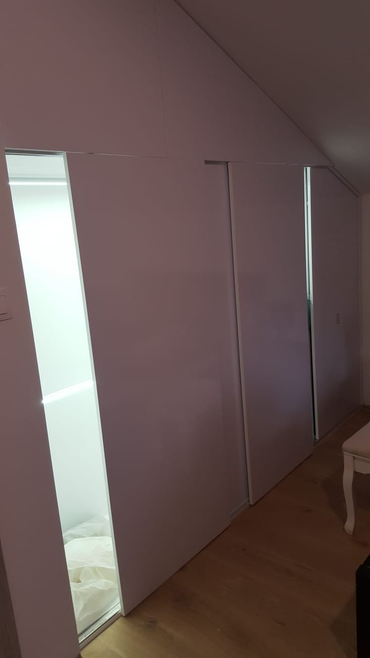 Biała szafa wnękowa z przesuwnymi drzwiami na poddaszu, widoczna zawartość jednej z półek i fragment drewnianej podłogi.