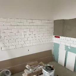 Ściana częściowo wyłożona białymi, prostokątnymi płytkami typu metro, widoczne narzędzia glazurnicze i wiaderka z zaprawą, fragment ściany przygotowany pod dalsze prace.