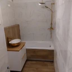 Nowoczesna łazienka z marmurowymi płytkami, złotą armaturą i drewnianymi akcentami, umywalka nablatowa w kolorze białym ze złotym rantem.