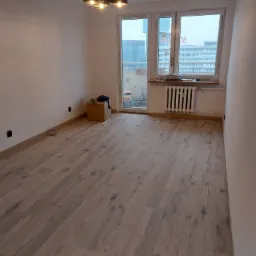 Pusty pok&oacute;j po położeniu paneli podłogowych w odcieniach brązu, widoczny grzejnik, okno z widokiem na miasto i karton z narzędziami w rogu.