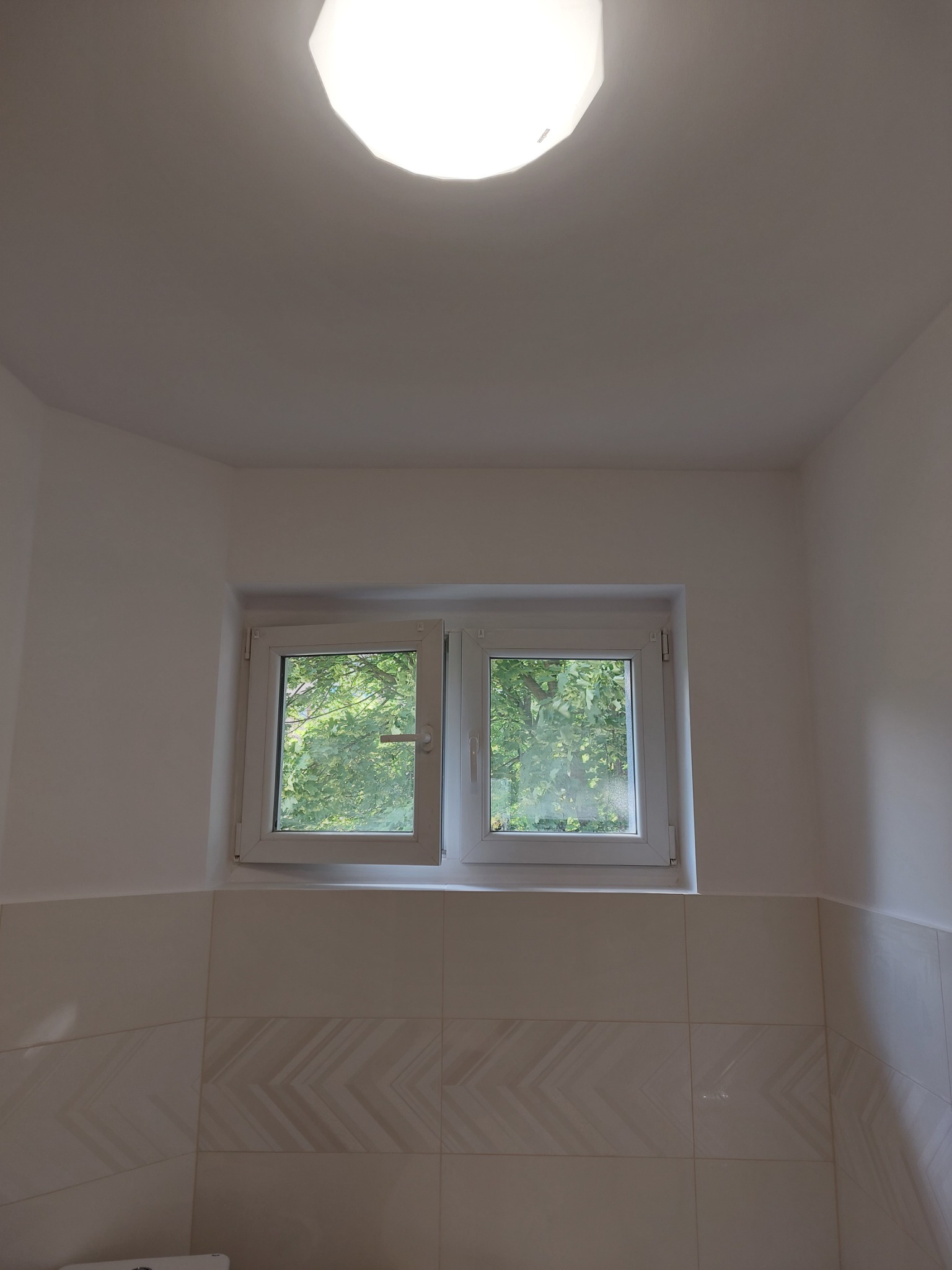 Biała łazienka po remoncie: widok na okno z zielenią za szybą, beżowe płytki z delikatnym wzorem na ścianie, oraz okrągła lampa sufitowa.