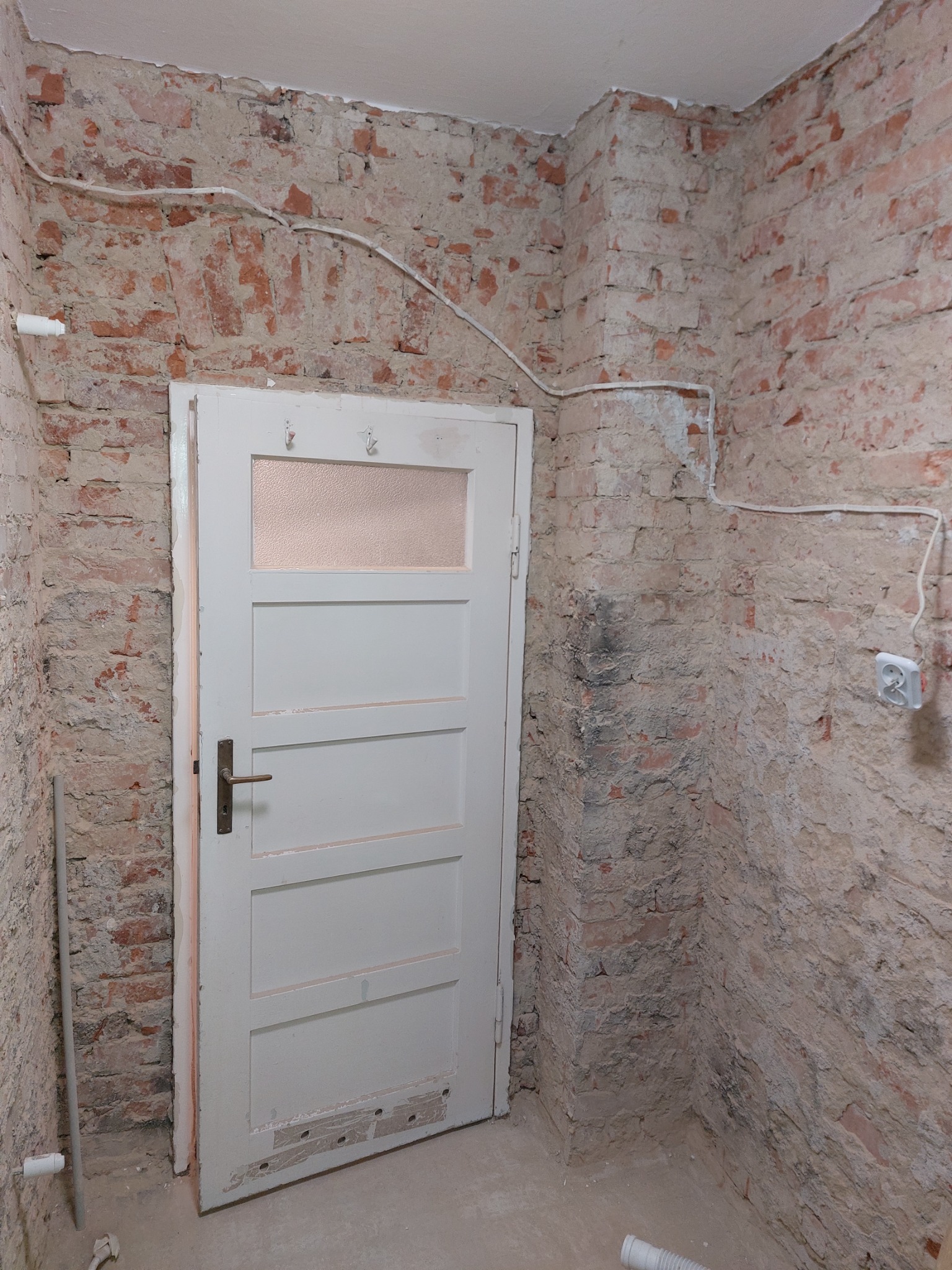 Surowa ściana z czerwonej cegły i białe drzwi z szybką w trakcie remontu, widoczne przewody elektryczne i gniazdko.
