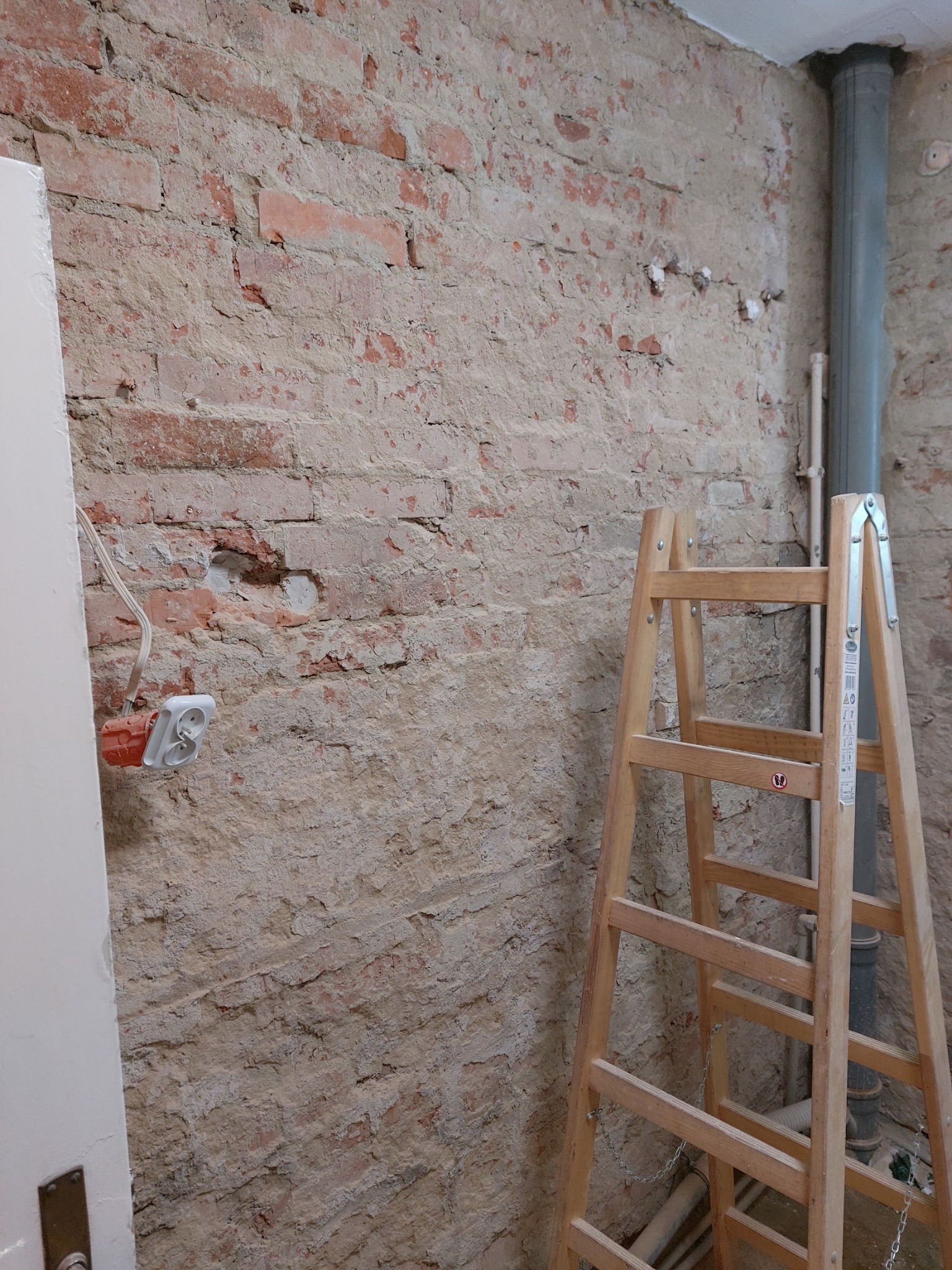 Ściana z cegły w trakcie przygotowania do prac wykończeniowych, widoczna drabina, gniazdko elektryczne i rura