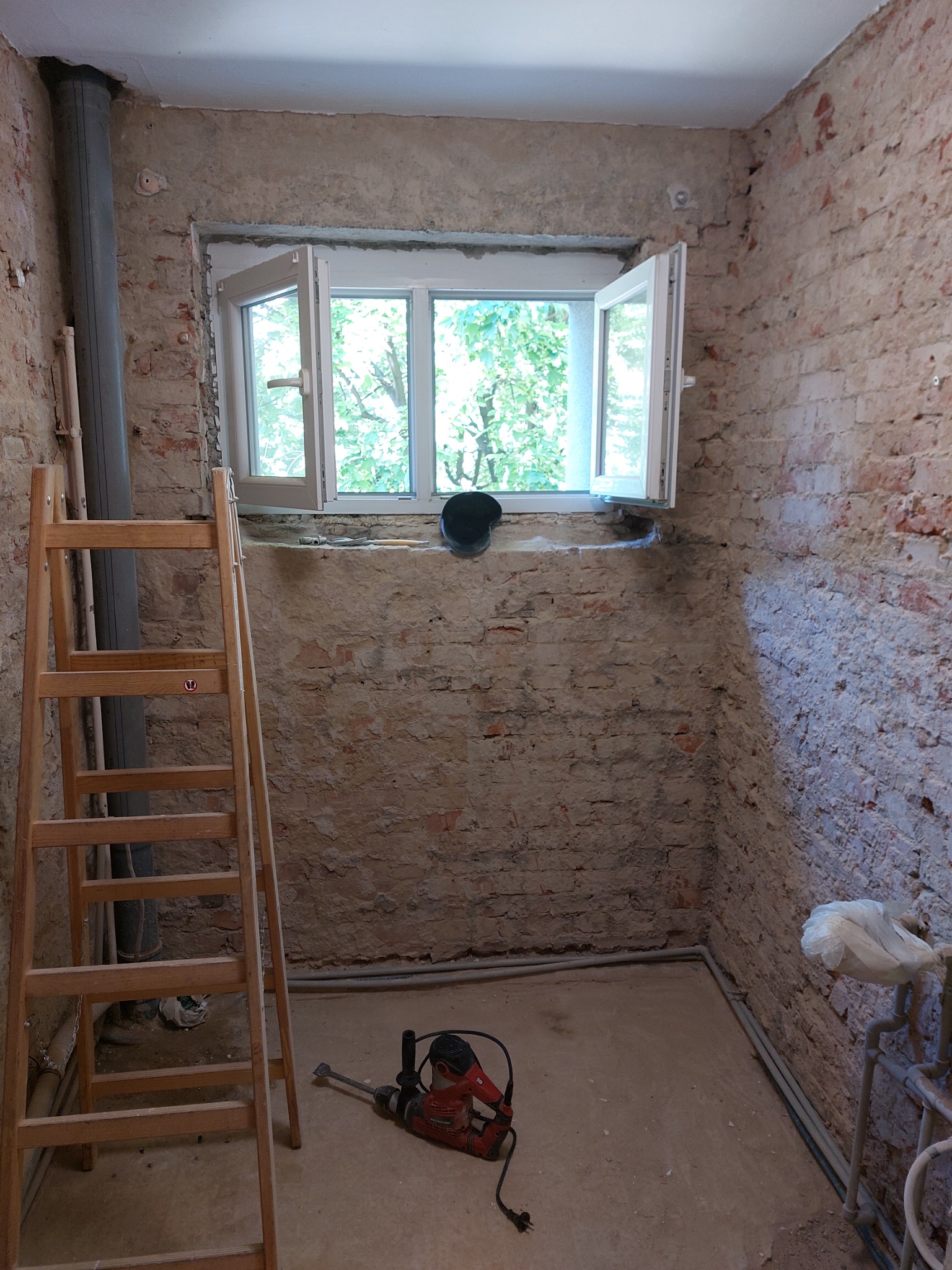 Pokój w trakcie prac przygotowawczych do remontu: odsłonięta cegła, drabina drewniana oparta o ścianę, wiertarka udarowa leżąca na podłodze, otwarte okno z widokiem na zieleń.