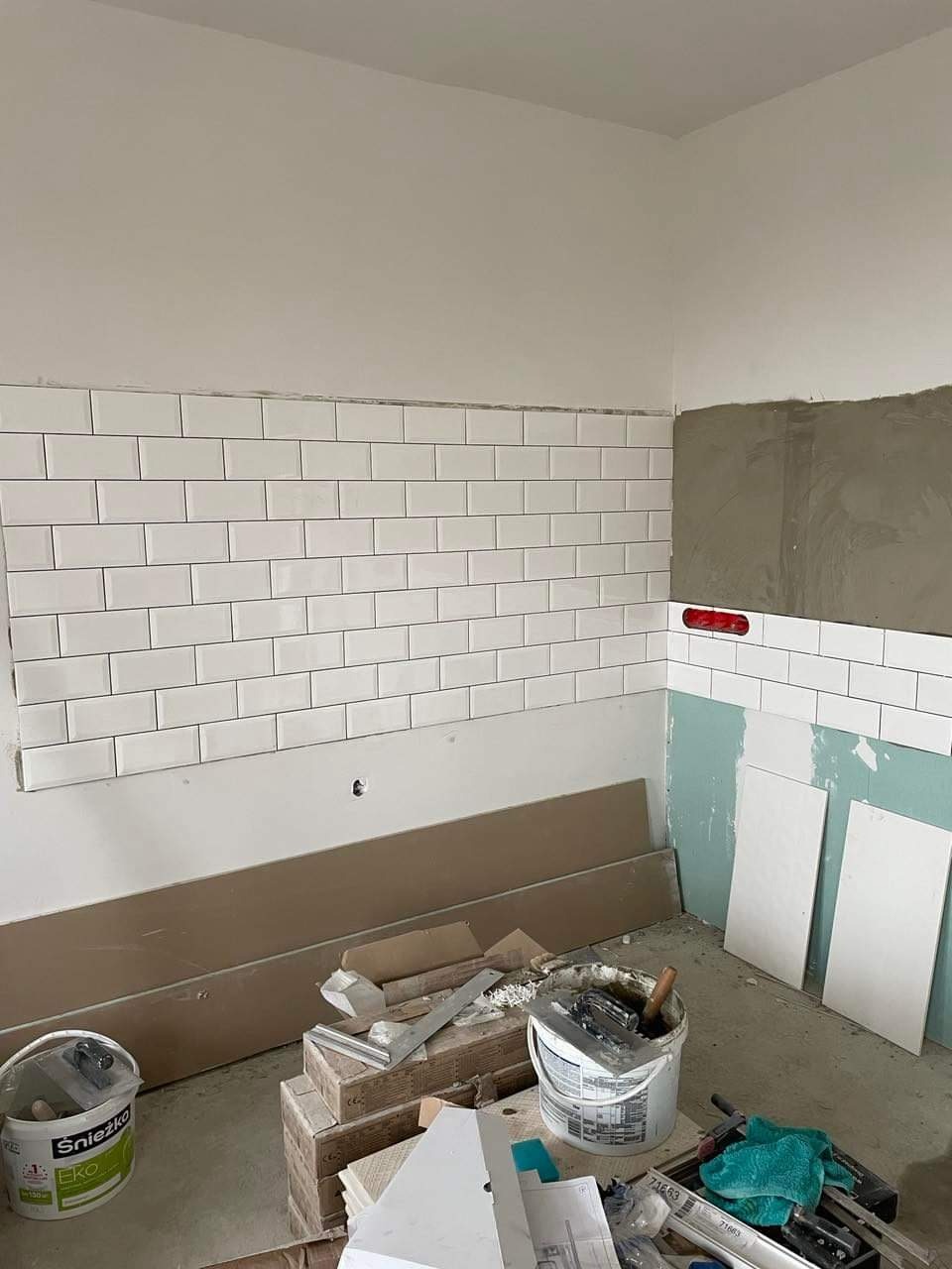 Ściana częściowo wyłożona białymi, prostokątnymi płytkami typu metro, widoczne narzędzia glazurnicze i wiaderka z zaprawą, fragment ściany przygotowany pod dalsze prace.