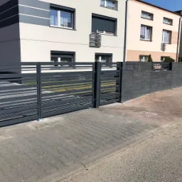 Nowoczesna, szara brama przesuwna z poziomymi panelami, dopasowana kolorystycznie do betonowego muru z wbudowaną skrzynką pocztową, na tle dwukondygnacyjnego domu.