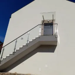 Zewnętrzne schody z szarą nawierzchnią prowadzące do drzwi wejściowych, zabezpieczone szklaną balustradą z metalowymi elementami mocującymi, nad drzwiami szklany daszek.