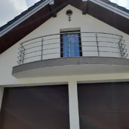 Biały dom z brązowymi bramami garażowymi i balkonem z metalową, minimalistyczną konstrukcją oraz oknem z niebieskimi zasłonami.