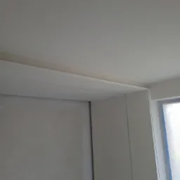 Ściana z nałożoną gładzią szpachlową, widoczne łączenie płyty gipsowej i fragment okna zabezpieczonego folią malarską.