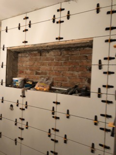 Ściana w trakcie układania białych płytek z użyciem krzyżyków dystansowych, widoczne fragmenty surowej cegły w otworze okiennym, wiadro z zaprawą i narzędzia w tle.