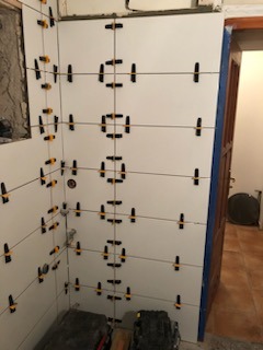 Świeżo ułożone, białe płytki ścienne w łazience, z zastosowaniem systemu poziomowania płytek (klipsy i kliny) na fugach, widoczne przewody instalacji wodnej i fragment futryny drzwi.