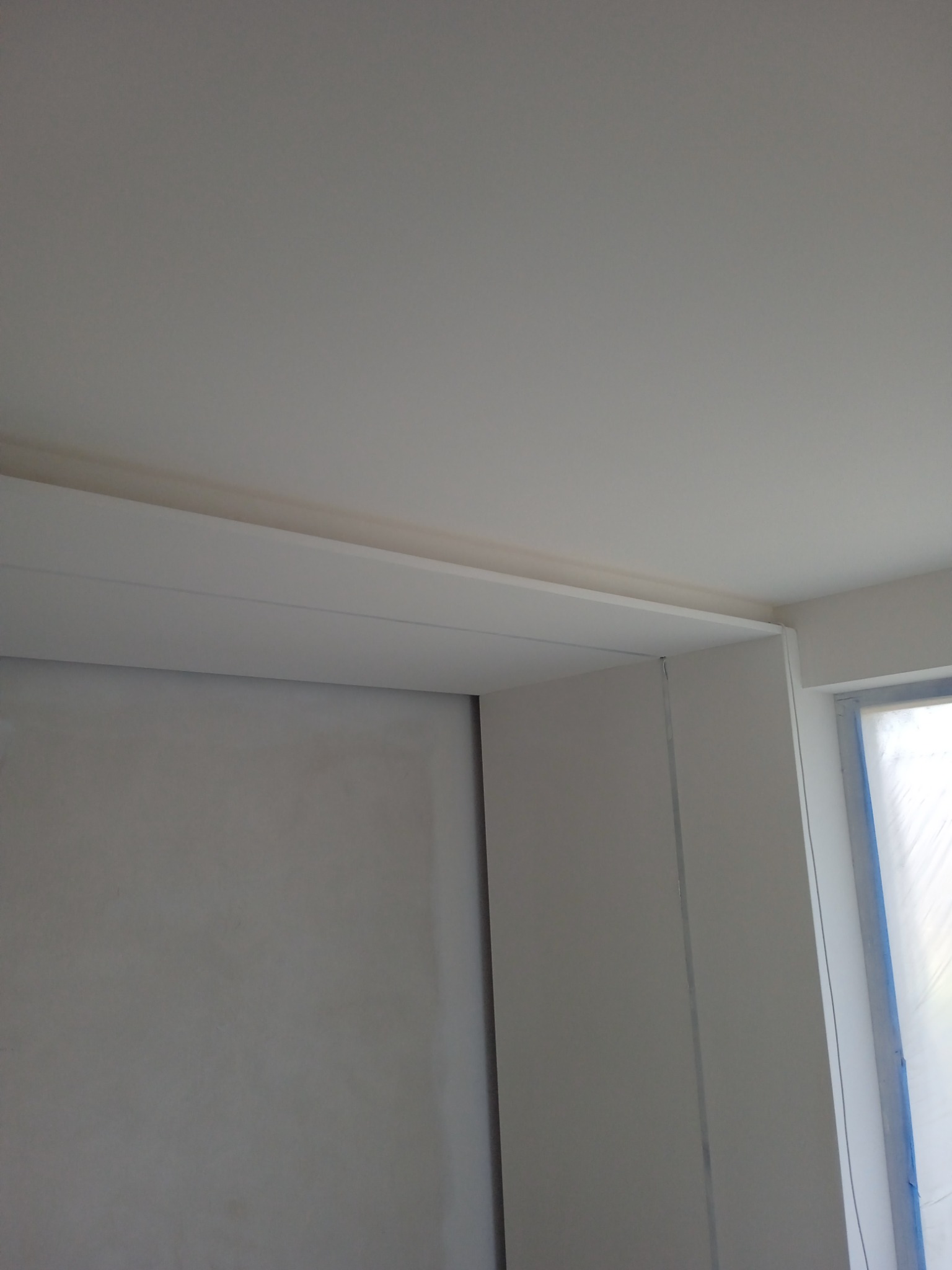 Narożnik ściany po nałożeniu gładzi, widoczny fragment okna zabezpieczonego folią malarską.