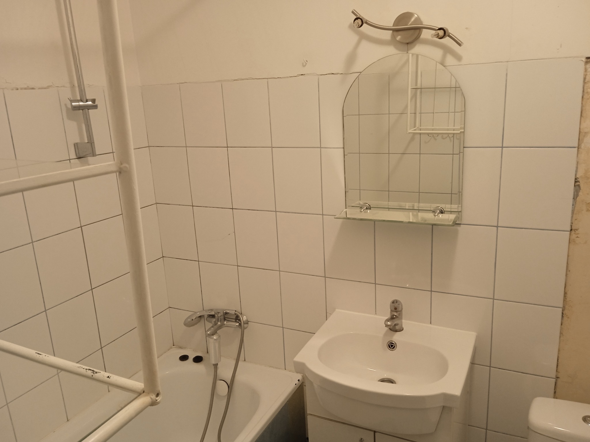 Łazienka przed remontem: białe kwadratowe płytki, wanna z metalową ramą, umywalka z lustrem i oświetleniem, widoczne uszkodzenia na ścianach.