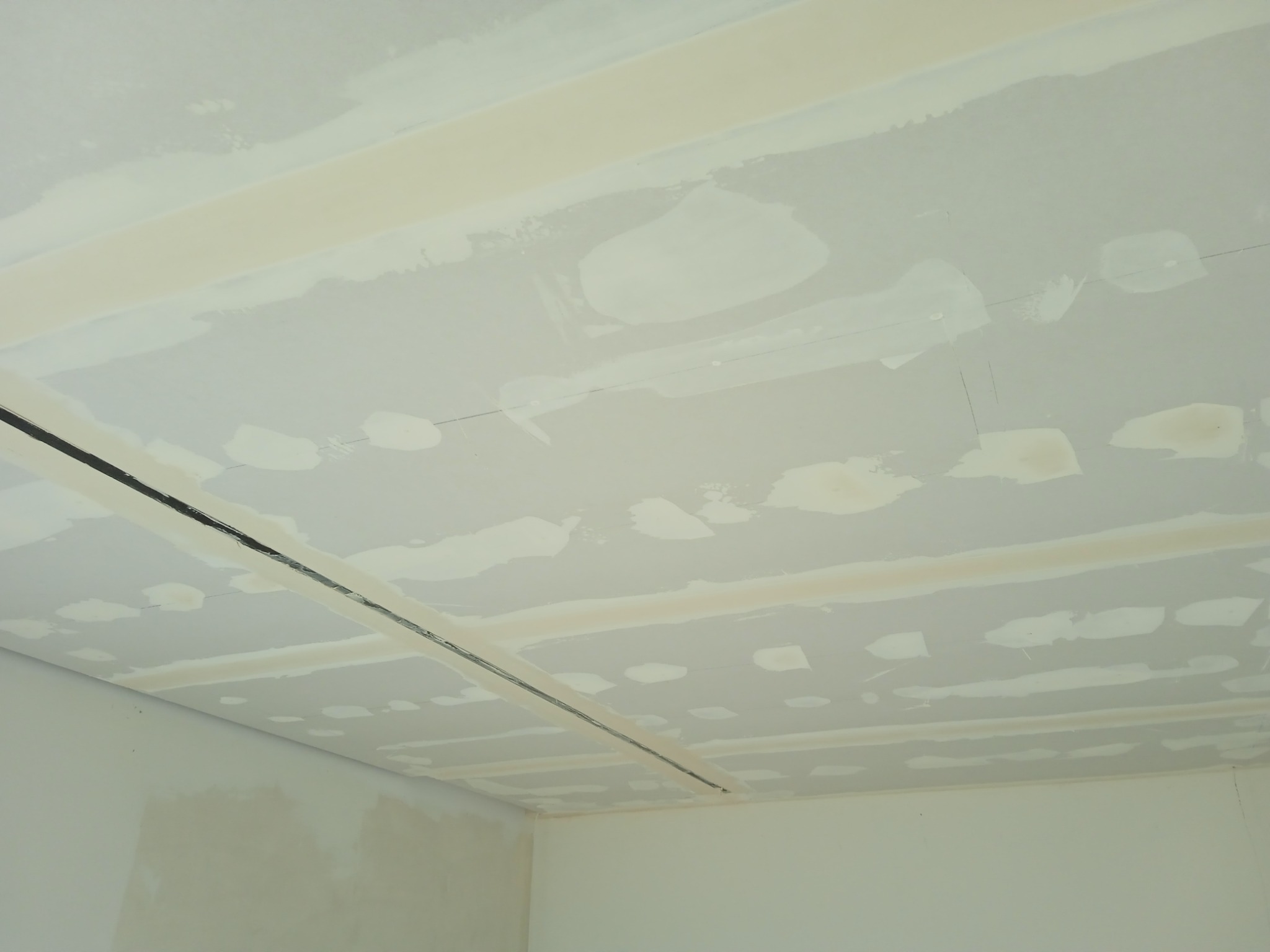 Surowy sufit i ściany po wstępnym nałożeniu gładzi, widoczne łączenia płyt kartonowo-gipsowych i plamy po szpachli, przygotowanie do dalszych prac wykończeniowych.