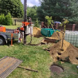 Mobilna wiertnica na przyczepie w trakcie prac ziemnych, widoczne wykopane doły, pryzma piasku, zbiorniki na wodę i rury w ogrodzie.