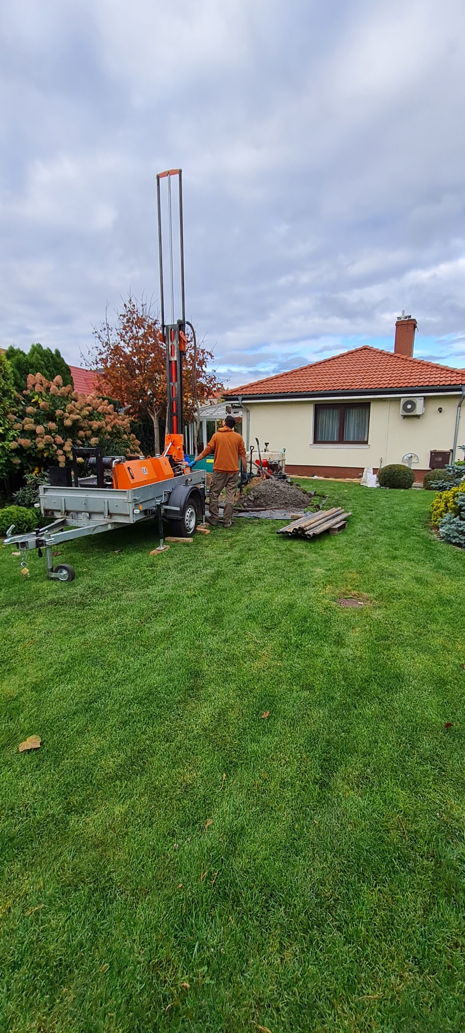 Pracownik w pomarańczowej bluzie obsługuje wiertnicę zamontowaną na przyczepie podczas wiercenia studni na trawniku przed domem z czerwoną dachówką.