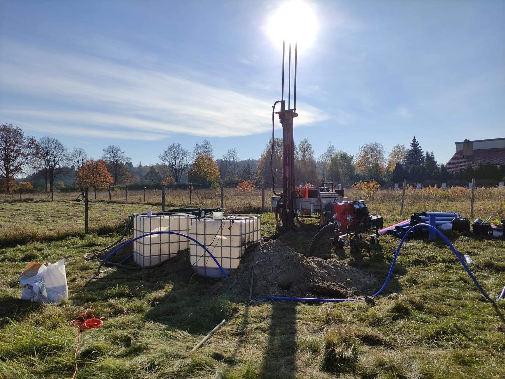 Wiertnica do studni na zewnątrz, obok dwa białe zbiorniki na wodę, w tle drzewa i niebo z jasnym słońcem.