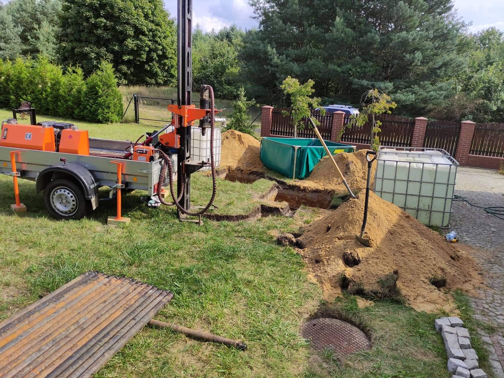 Mobilna wiertnica na przyczepie w trakcie prac ziemnych, widoczne wykopane doły, pryzma piasku, zbiorniki na wodę i rury w ogrodzie.