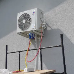 Biała jednostka zewnętrzna klimatyzacji marki Gree zamontowana na szarej ścianie budynku, podłączona do manometrów serwisowych za pomocą czerwonego i żółtego węża, widoczne rusztowanie i narzędzia...