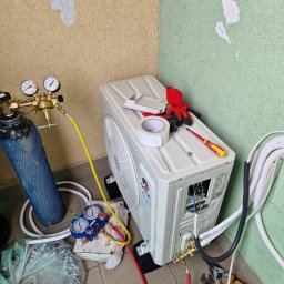 Klima Partner - Montaż klimatyzatora: butla z gazem, manometry, narzędzia, przewody i rękawice obok jednostki zewnętrznej na balkonie z zielono-beżową ścianą.