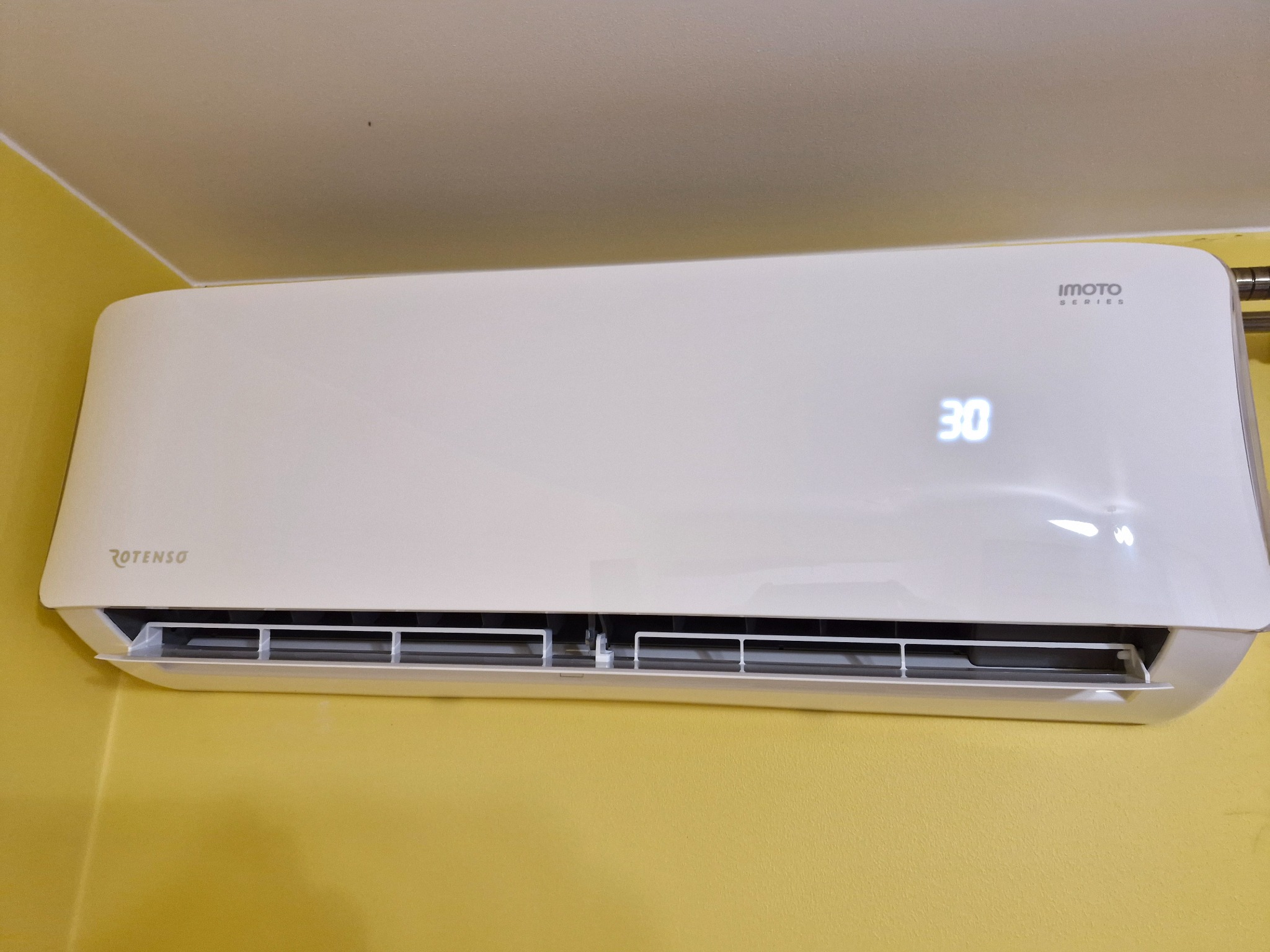 Biały klimatyzator Rotenso Imoto Series zamontowany na żółtej ścianie, wyświetlający temperaturę 30 stopni.