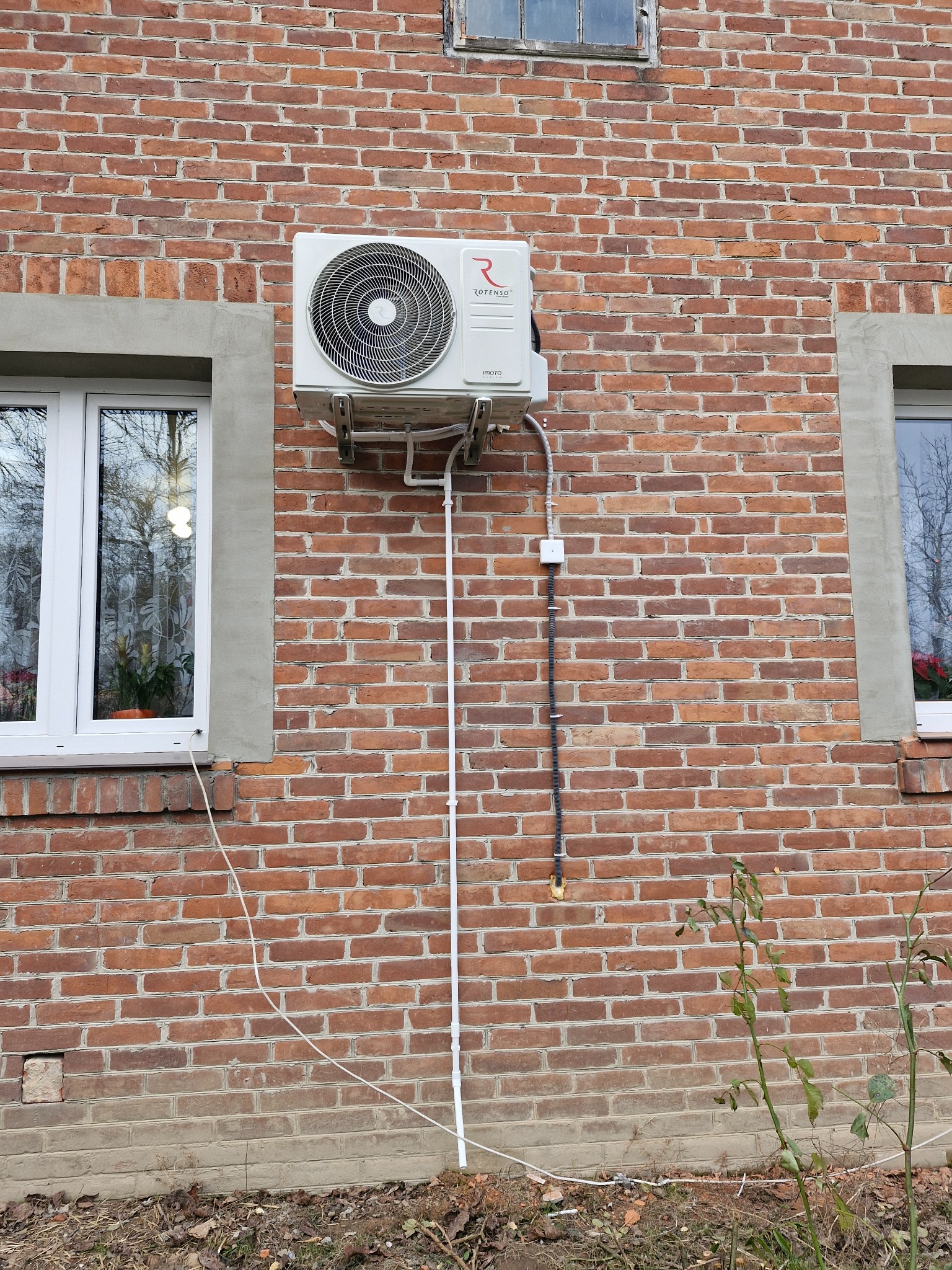 Zewnętrzna jednostka klimatyzacji zamontowana na ceglanej ścianie domu jednorodzinnego, widoczne przewody i instalacja elektryczna.