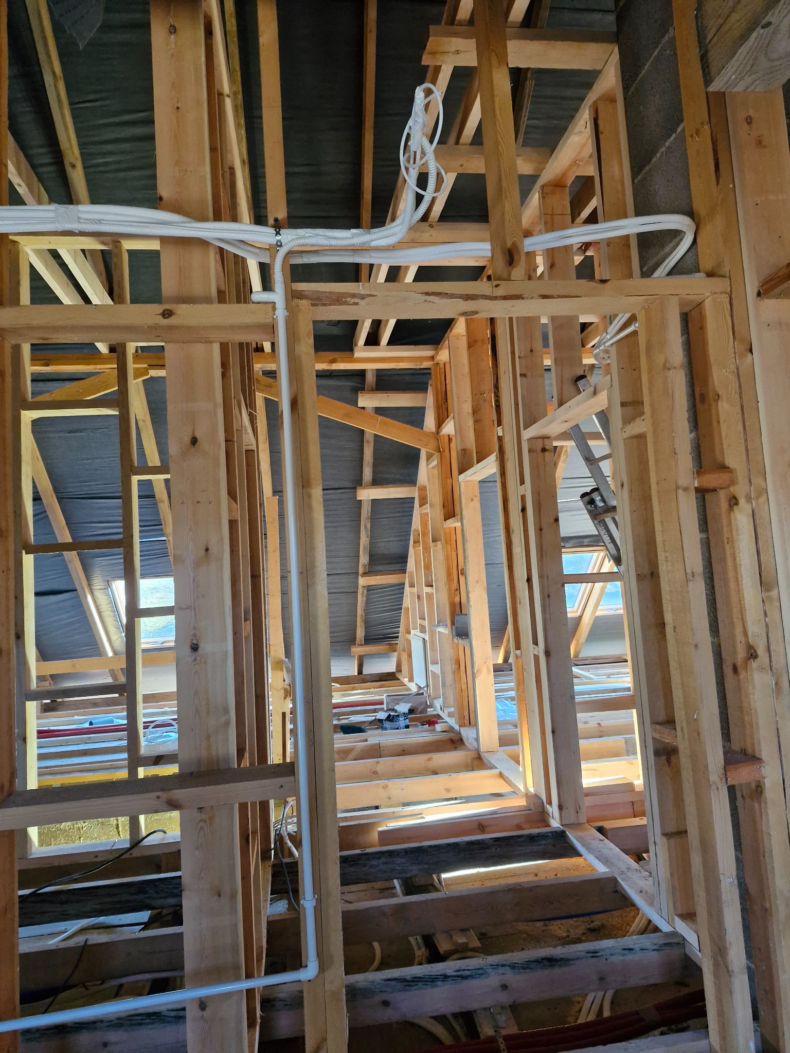 Surowa konstrukcja drewniana z białymi rurami instalacji pod sufitem w trakcie budowy domu, widoczne elementy izolacji i rusztowanie.