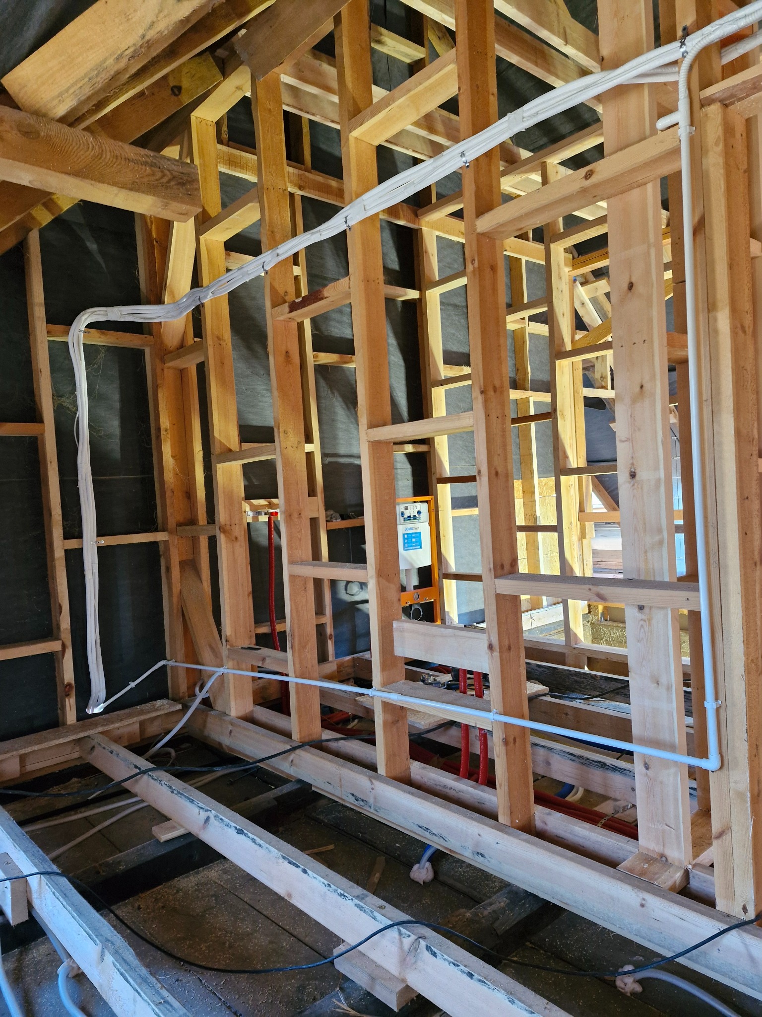 Wnętrze w trakcie budowy z drewnianą konstrukcją szkieletową, widoczne białe rury instalacyjne oraz przewody elektryczne prowadzone wzdłuż ścian, w tle czarna folia izolacyjna.